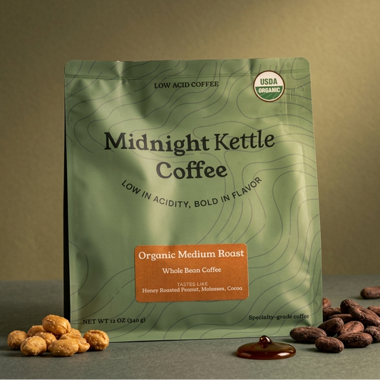 A stand-up pouch of Midnight Kettle Coffee, Organic Medium Roast, displayed on a dark gray surface with coffee beans and a dollop of coffee around it. The pouch is sage green with black text and a small orange label.