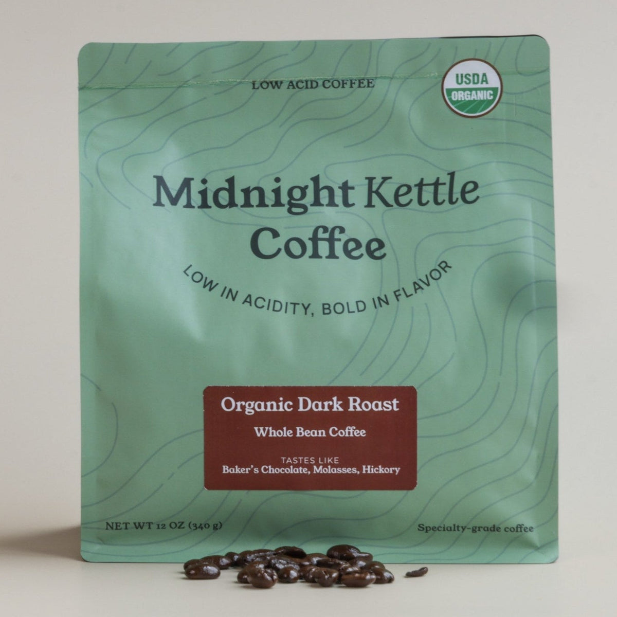 A pouch of Midnight Kettle Coffee Organic Dark Roast stands upright on a light beige background with a few coffee beans scattered in front. The pouch is mint green with dark green topographic contour lines, black text, and a dark red label near the bottom.