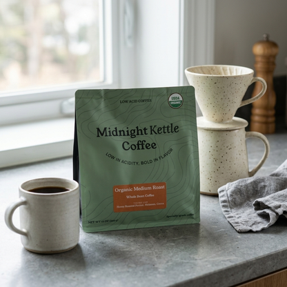 A package of Midnight Kettle Coffee stands on a kitchen counter beside a cup of black coffee, stacked ceramic mugs, a cloth, and a window.